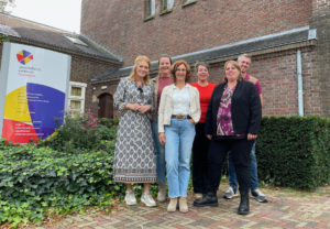 V.l.n.r.: Hilde van Bree, Kim van Sinten, Janneke Bosboom, Thea Reinders, Silvia Manders en Merijn Godefrooij. Ze staan voor een stenen gebouw, links in beeld het bord van de huisartsenpraktijk.